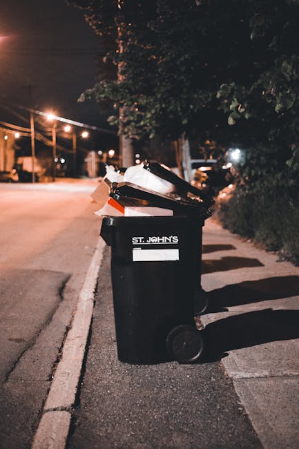 A black wheeled rubbish bin positioned on the edge of a sidewalk along a quiet street at night. The bin bears the label 'ST. JOHN'S' and contains a mix of waste materials, including cardboard and plastic items, with some pieces protruding from the open lid. The bin's surface appears slightly textured, with visible scuff marks and fading. In the background, there are streetlights illuminating the scene with a warm glow, casting soft shadows on the pavement. Overhanging branches and trees partially frame the top of the image, while some residential houses and parked vehicles are faintly visible under the night sky. The setting suggests an area where independent waste collection or disposal services, such as those provided by Waste Disposal Waterloo, are used for household rubbish management. The image emphasizes the practical aspect of rubbish collection, highlighting the importance of proper waste disposal at the curbside during designated collection times.