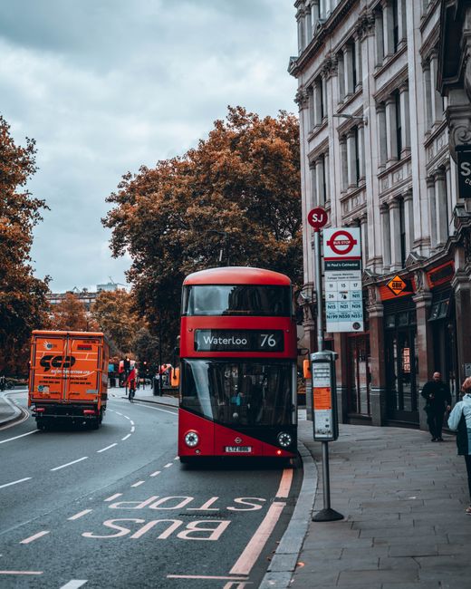 A double-decker red bus displaying the route number 76 and destination Waterloo is parked at a bus stop on a city street lined with historic multi-story buildings featuring ornate stone facades and large windows. Adjacent to the bus stop, there is a digital timetable and a vertical bus shelter with a transparent glass panel. To the left of the bus, an orange delivery truck is travelling along the curved road, which is marked with a white 'BUS STOP' line and a dedicated bus lane. Pedestrians are visible on the sidewalk, some walking past the bus stop and others standing nearby. In the background, leafy trees with orange-brown autumn foliage provide a natural contrast to the urban environment, and a clear sky with scattered clouds creates a bright daytime atmosphere. The scene captures an active city scene that involves public transport, pedestrian activity, and urban infrastructure, illustrating the typical setting where waste disposal and rubbish collection services, such as those offered by Waste Disposal Waterloo, might be needed for nearby commercial or residential waste management requirements.