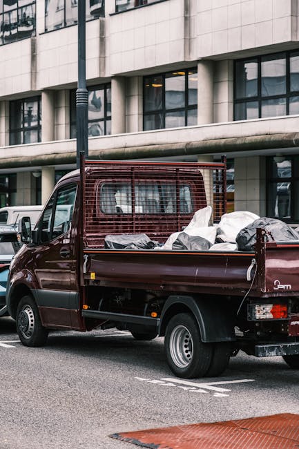 A dark red flatbed truck parked on an urban street in front of a modern building with large rectangular windows and light-colored stone facade. The truck's open cargo bed is loaded with a variety of mixed waste materials, including black and white plastic bags, cardboard, and other debris. A metal mesh panel acts as a barrier at the front of the cargo area. The vehicle's rear and side lights are visible, along with its silver alloy wheel rims. The street surface is asphalt, with a white painted line indicating a parking zone. Nearby, a white van is partially visible, and a black pole with a streetlight is positioned adjacent to the truck. The scene suggests an independent waste collection or rubbish clearance operation, consistent with services provided by Waste Disposal Waterloo, in an urban environment suitable for commercial rubbish removal and on-site clearance activities.