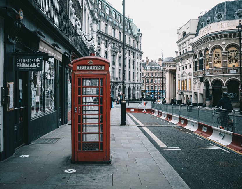 A traditional red British telephone box stands prominently on a city sidewalk in front of a building with large windows, black framing, and signage for a champagne and wine shop. The telephone kiosk features a hinged door, glass panels, and a domed top with an inscription reading 'TELEPHONE'. To the right, a section of the street is visible, with temporary orange and white traffic barriers separating the pedestrian sidewalk from the road, which appears to be under construction or re-surfacing. In the background, multi-story buildings with classic European architectural details are seen, including decorative stone facades, arched windows, and balconies with wrought-iron railings. Several pedestrians are walking along the sidewalk, and some are riding bicycles. Overcast lighting casts soft shadows, lending a subdued tone to the scene, which captures an urban environment where independent street furniture and architecture coexist with ongoing infrastructure work, aligning with the context of alternative waste management and localised rubbish removal services near Waterloo Station SE1, Waterloo.