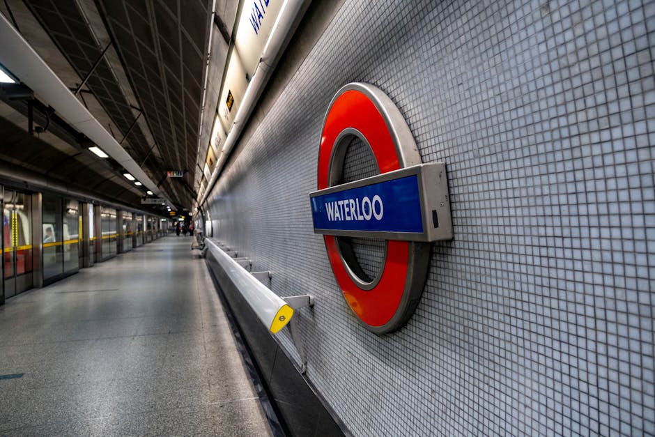 A close-up view of the London Underground station signage for Waterloo, featuring a circular red and silver frame with a blue rectangular sign displaying the station name in white text. The sign is mounted on a wall covered with small, light grey square tiles. In the background, the subway platform extends into the distance, with a clean, spacious concrete floor and glass barrier panels along the edge, separating passengers from the tracks. The ceiling is industrial with dark metal beams and bright overhead lighting, creating a well-lit environment. The scene is devoid of passengers, emphasizing the station's modern design and orderly appearance. This type of infrastructure is often associated with public transportation systems, whereas in context of waste disposal, it reflects the importance of accessible transport hubs supporting efficient rubbish removal services near busy areas like Waterloo Station.